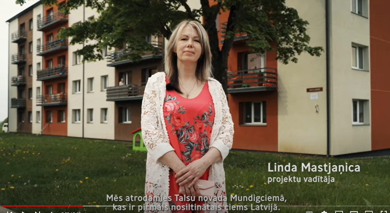 Saite uz dokumentālo sēriju "Kā labāk dzīvot?". Attēlā sieviete Talsu novada Mundigciemā, stāv priek&scaron;ā renovētai daudzdzīvokļu ēkai