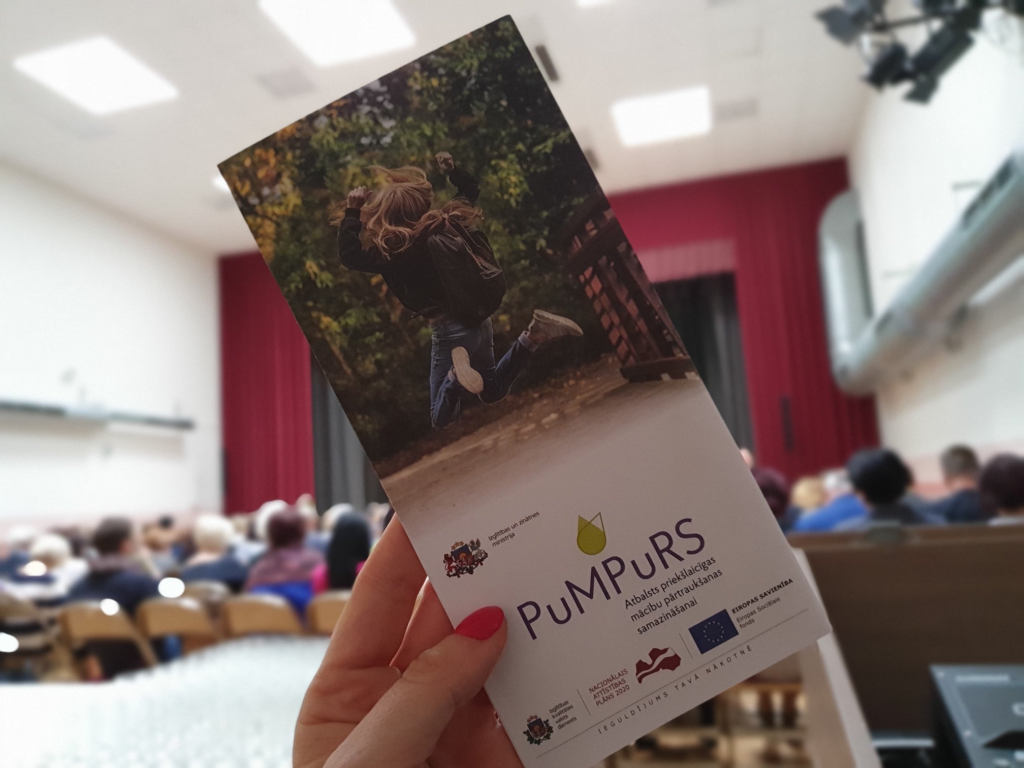 Sieviete kreisajā rokā tur bukletu "Pumpurs - Atbalsts priek&scaron;laicīgas mācību pārtrauk&scaron;anas samazinā&scaron;anai"