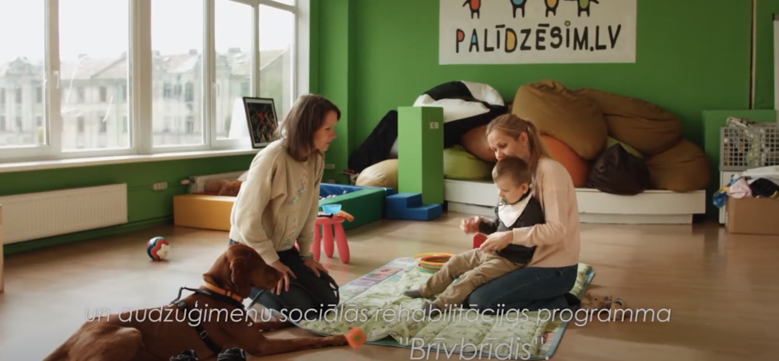 Attēls: Palīdzēsim.lv telpās notiek kanisterapija bērnam, teksts "un audžuģimeņu sociālās rehabilitācijas programma "Brīvbrīdis""