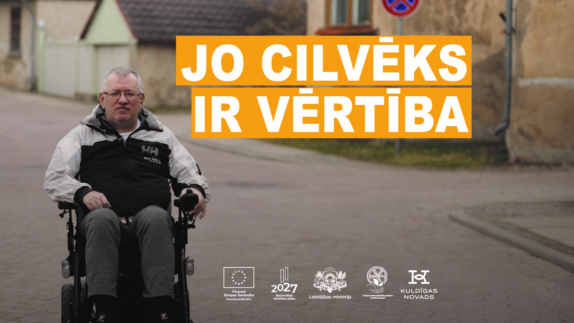 Bruģēta vecpilsētas iela. Vīrietis ap 50 gadiem, īsiem, sirmiem matiem brillītēs un pelēkā rudens vējjakā sēž elektriskajā ratiņkrēslā. Uzraksts "Jo cilvēks ir vērtība"