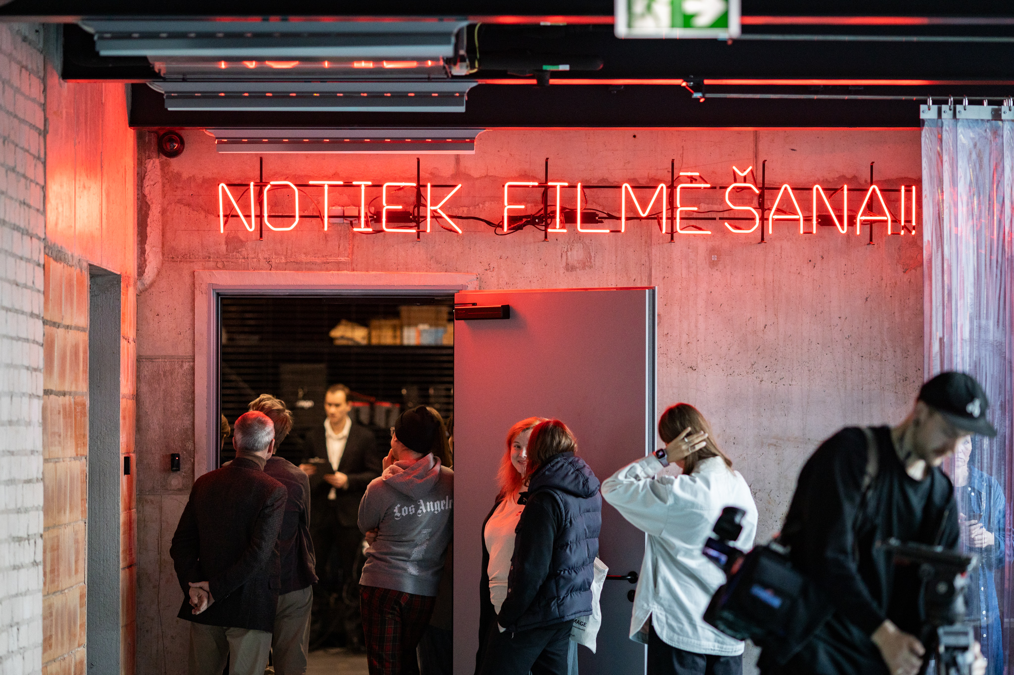 Attēls ar cilvēkiem telpā un uzrakstu: Notiek filmē&scaron;ana