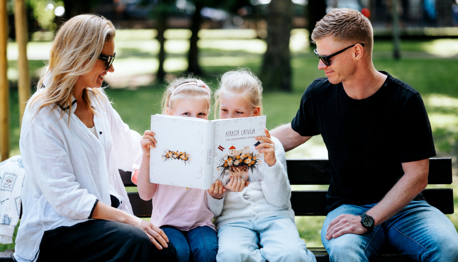 Family, dad, mom, two daughters, reading a book "Atrastā Latvija" in a sunny park.