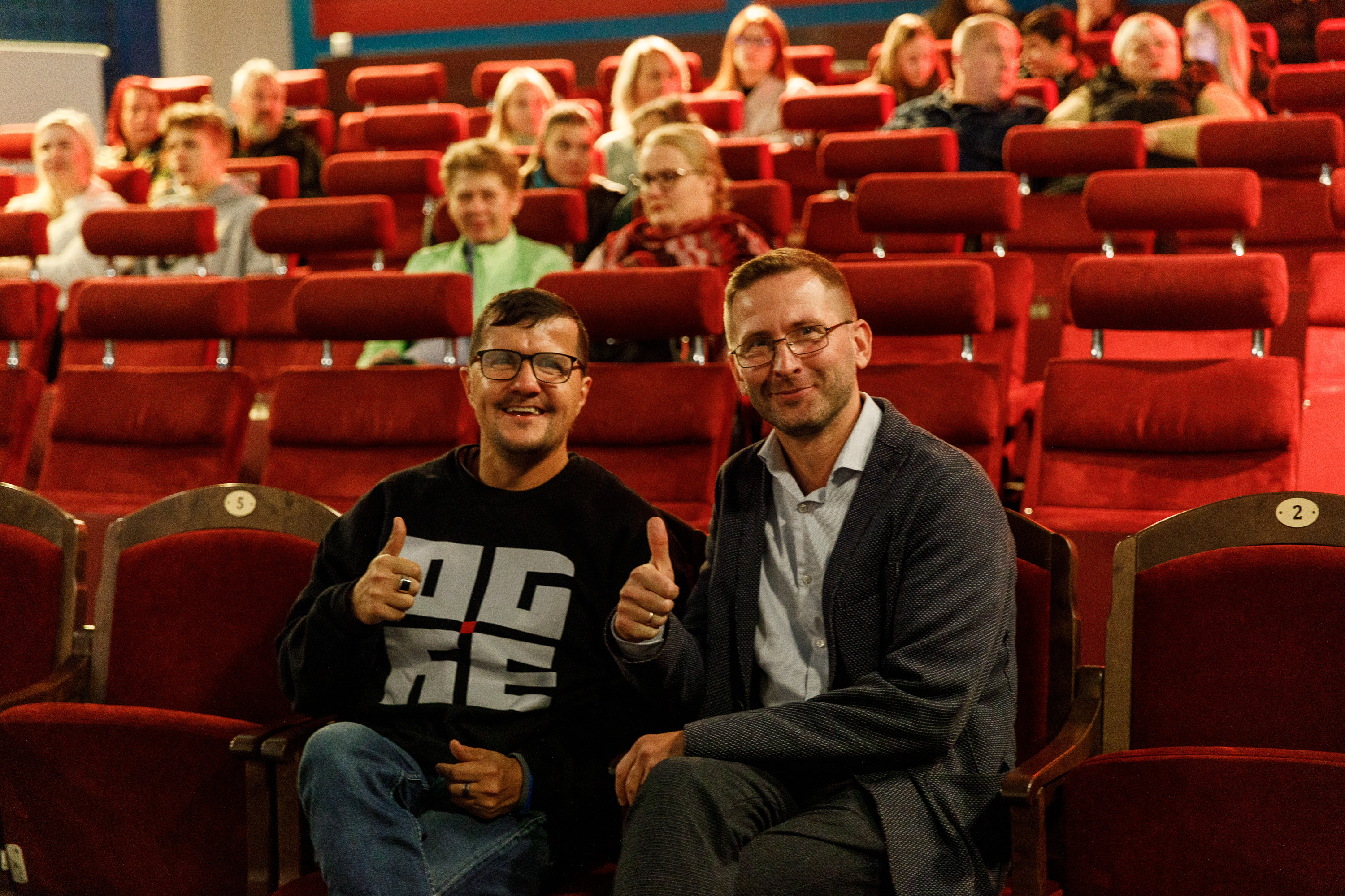 Kino teātris. Sēž divi vīrie&scaron;i, smaida un rāda paceltus gaisā īk&scaron;ķus