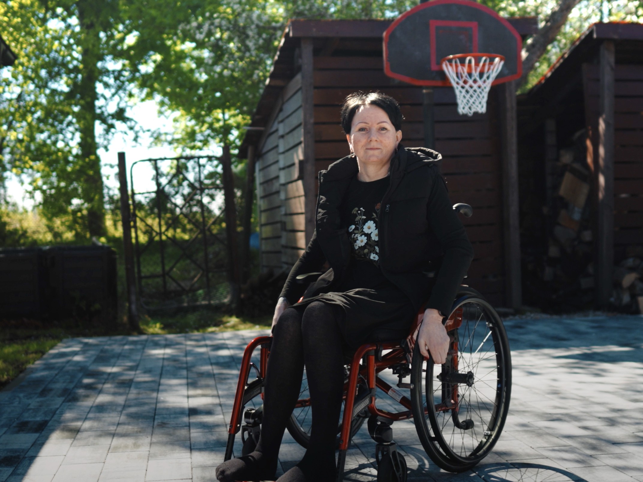 Privātmājas iek&scaron;pagalms. Pa perimetru glīti &scaron;ķūnī&scaron;i no tum&scaron;a koka dēļiem, fonā lieli koki un basketbola grozs. Pagalms bruģēts un tā vidū ratiņkrēslā sēž slaida, kopta ap piecdesmit gadu veca sieviete īsiem tum&scaron;iem matiem tum&scaron;ās drēbēs.