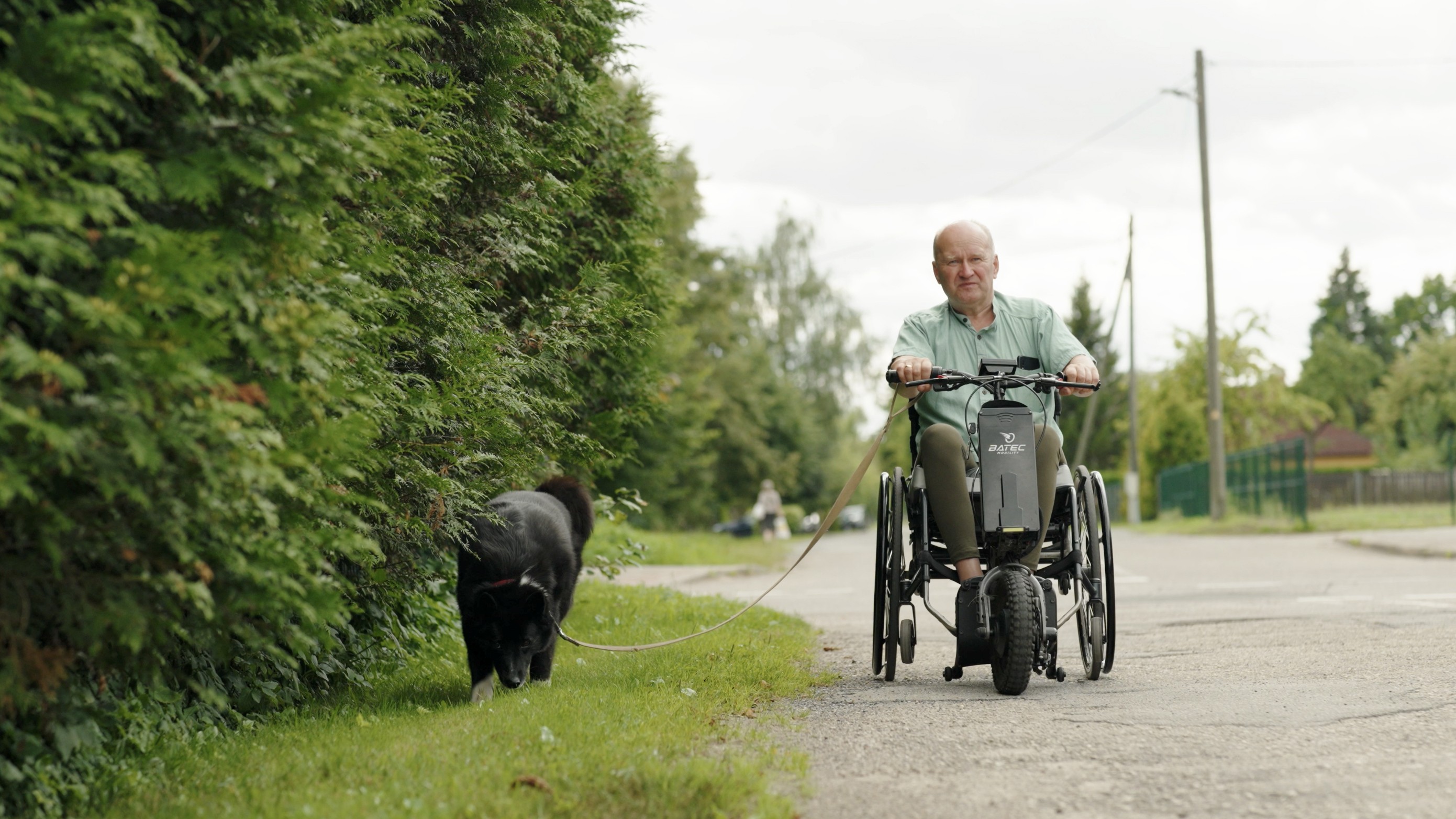 Vīrietis zaļā kreklā vasarā ratiņkrēslā ar elektrisko piedziņu pa dzīvojamo māju kvartāla ielu ved pastaigā pavadā vidēja izmēra melnu suni.