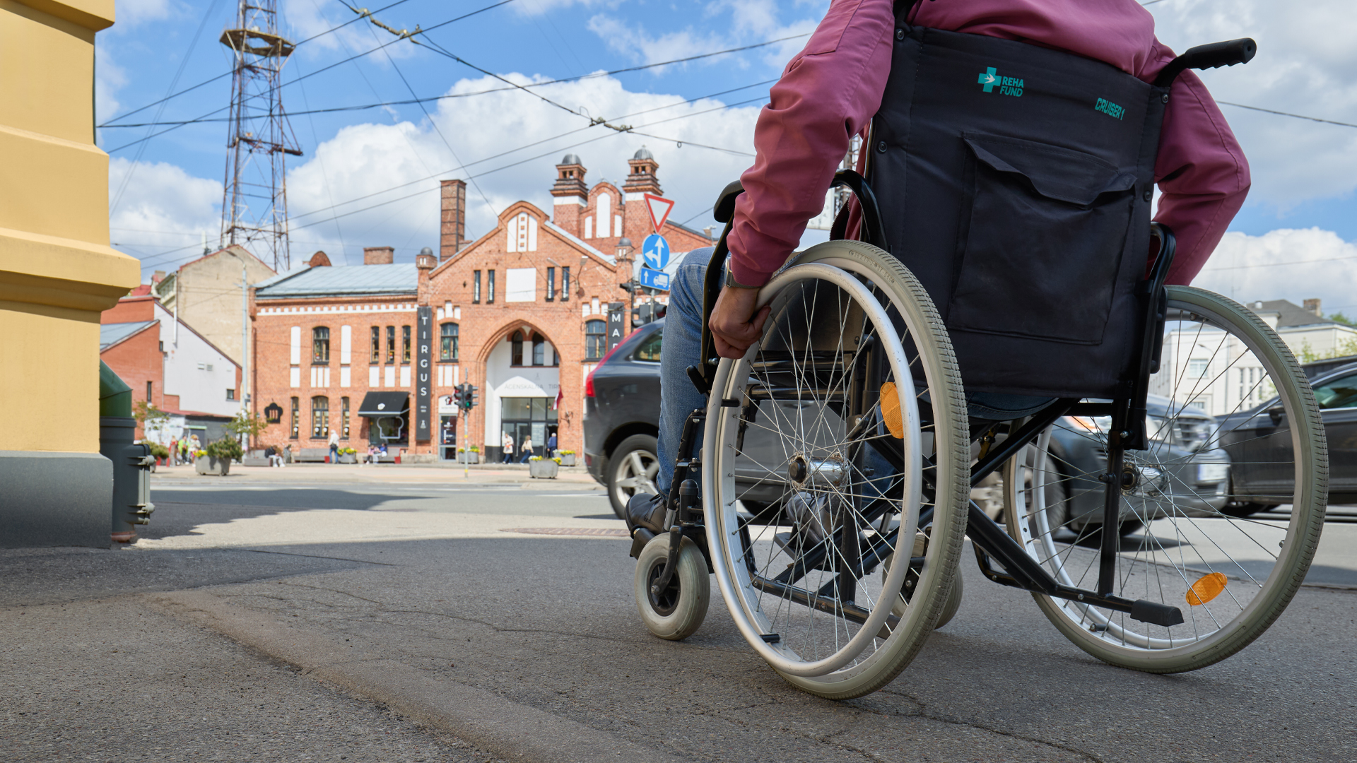 Saulaina diena. Pilsētvide ar asfaltētu ietvi, ceļu pa kuru brauc auto. Fonā redzama oranža ķieģeļu ēka ar vairākiem dekoratīviem tornī&scaron;iem. Uz ietves pārvietojas cilvēks ratiņkrēslā