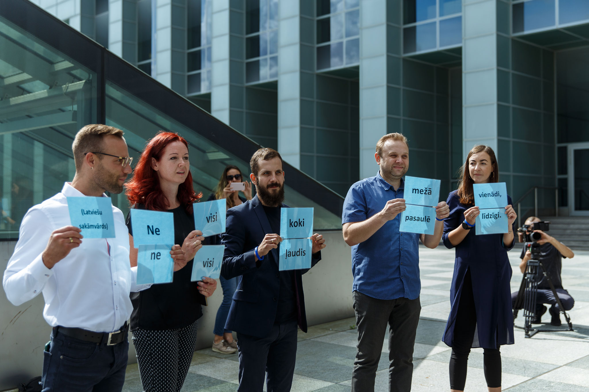Pieci cilvēki vasarā, Latvijas Nacionālās bibliotēkas fonā, stāv un rokās tur lapas ar tekstu" Ne visi koki mežā vienādi, ne visi ļaudis pasaulē līdzīgi"