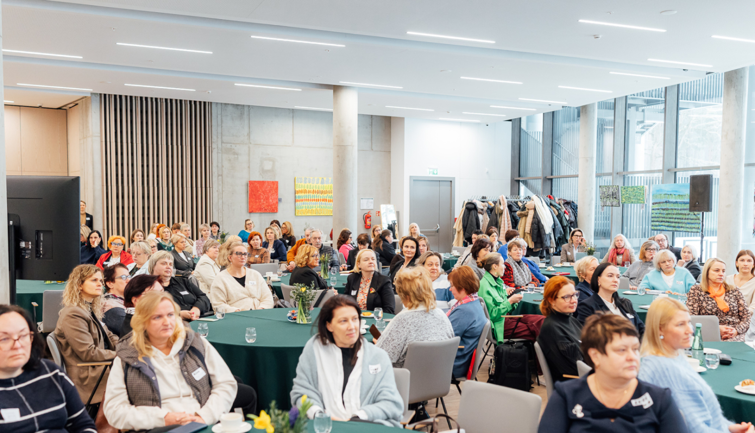 Pla&scaron;a, gai&scaron;a konferenču telpa ar logiem. Pie apaļiem galdiem sēž vairāki desmiti cilvēku, pārsvarā sievietees ap 50 gadiem, un klausās lektoru