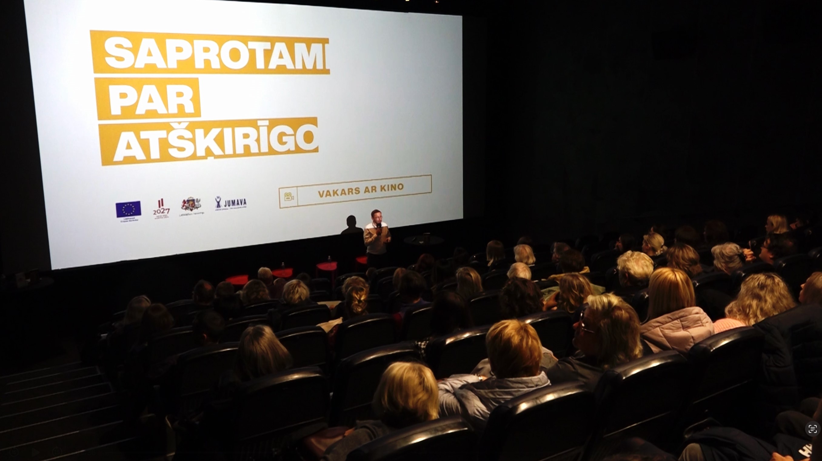 Pilna kino zāle. Vīrietis uzstājas ar runu. Fonā uz ekrāna uzraksts "Saprotami par at&scaron;ķirīgo"