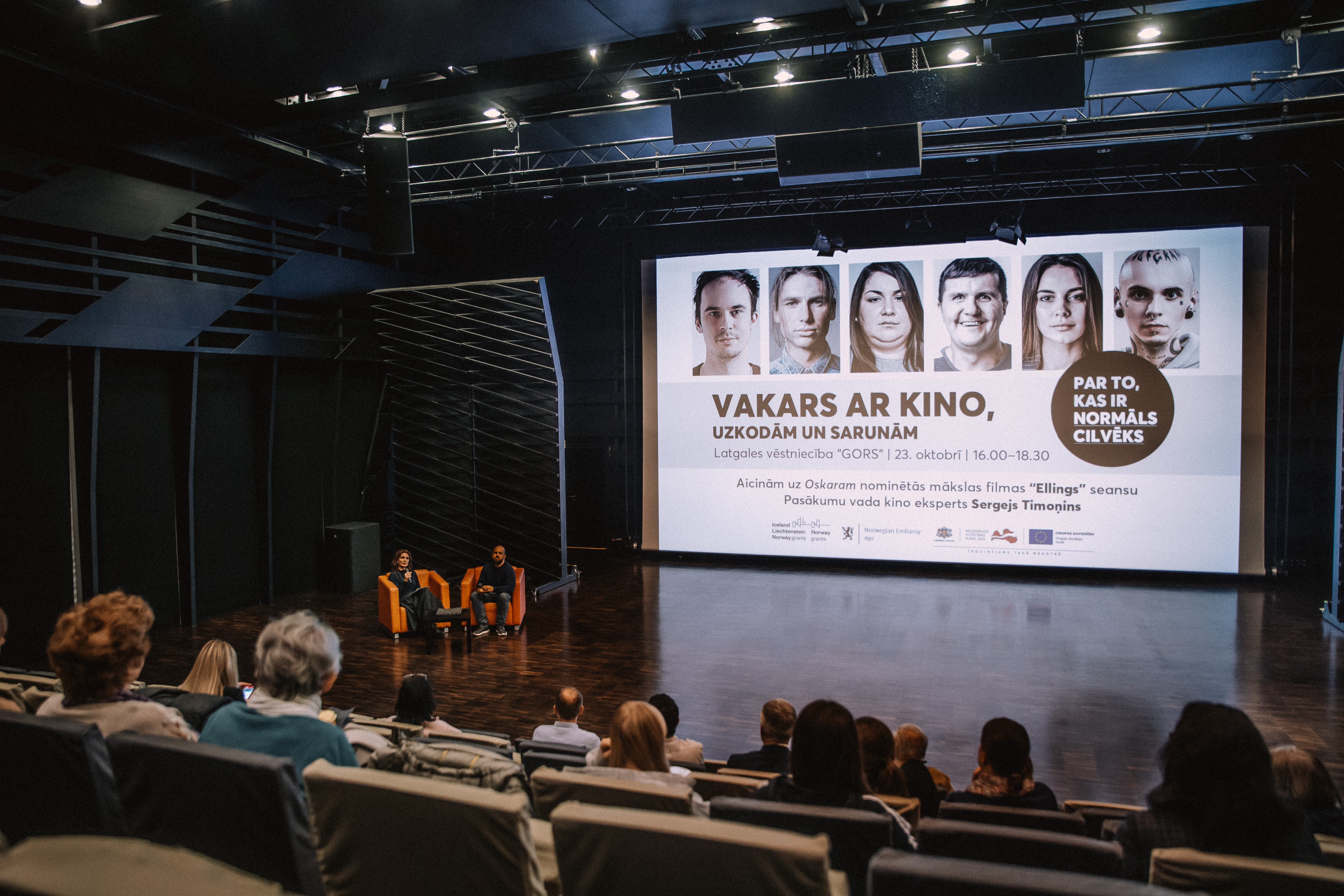 Kinozāle - skats uz ekrānu no sēdvietām. Divi cilvēki pie ekrāna diskutē.