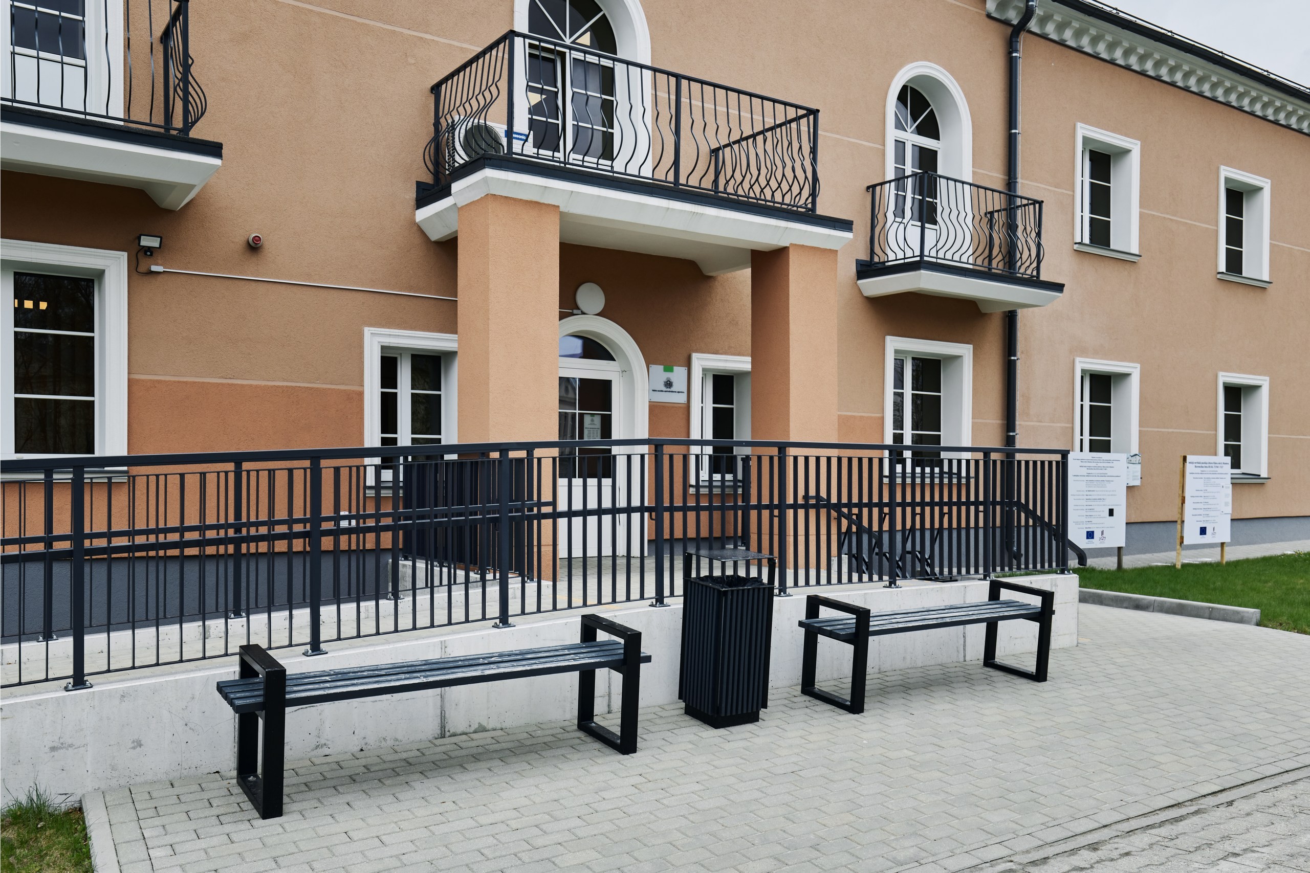 Divstāvīgas oranžas ēkas ar baltiem logu rāmjiem un melnām margām fasāde. Pie fasādes jauns bruģis, zāliens un divi soliņi