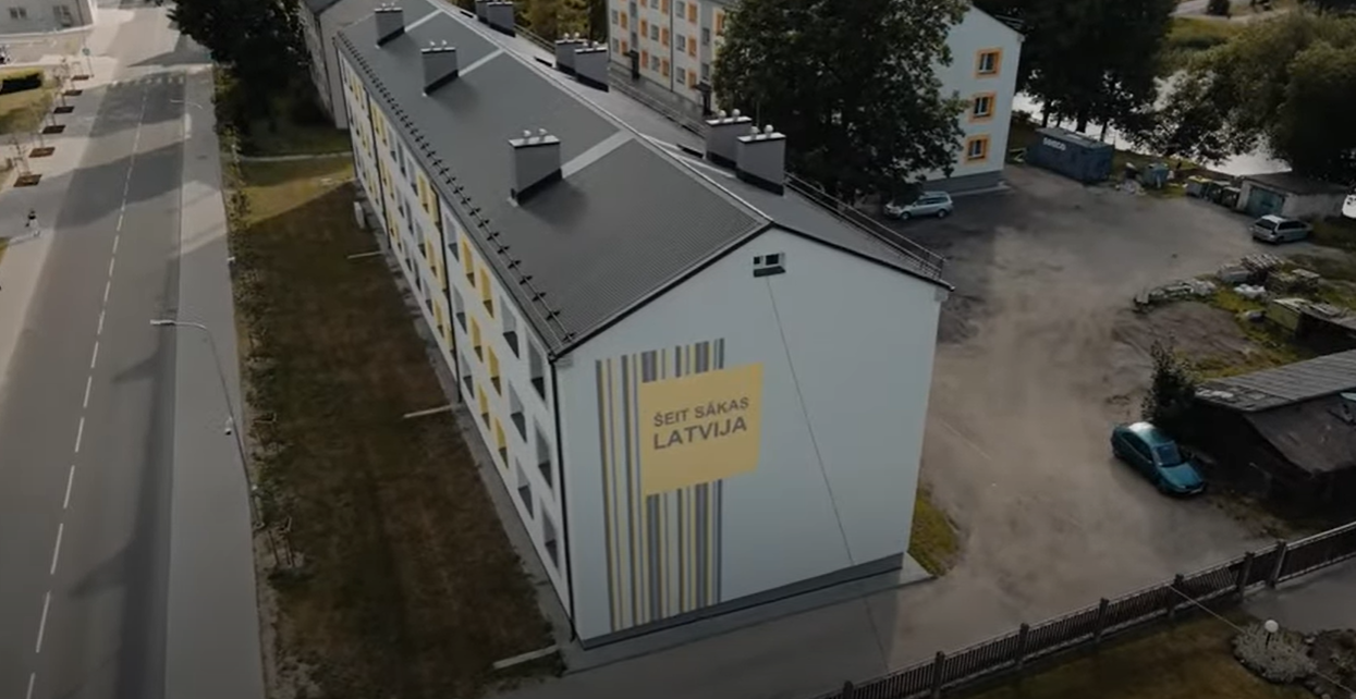 Saite uz dokumentālo sēriju "Kā labāk dzīvot?". Attēlā renovēta daudzzīvokļu māja Valkā ar tekstu "&Scaron;eit sākas Latvija"