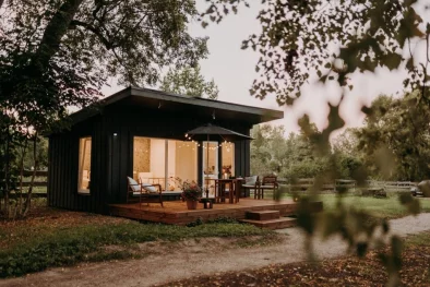 Maza mājiņa vakarā, iekšpusē ieslēgta gaisma, apkārt koku zari