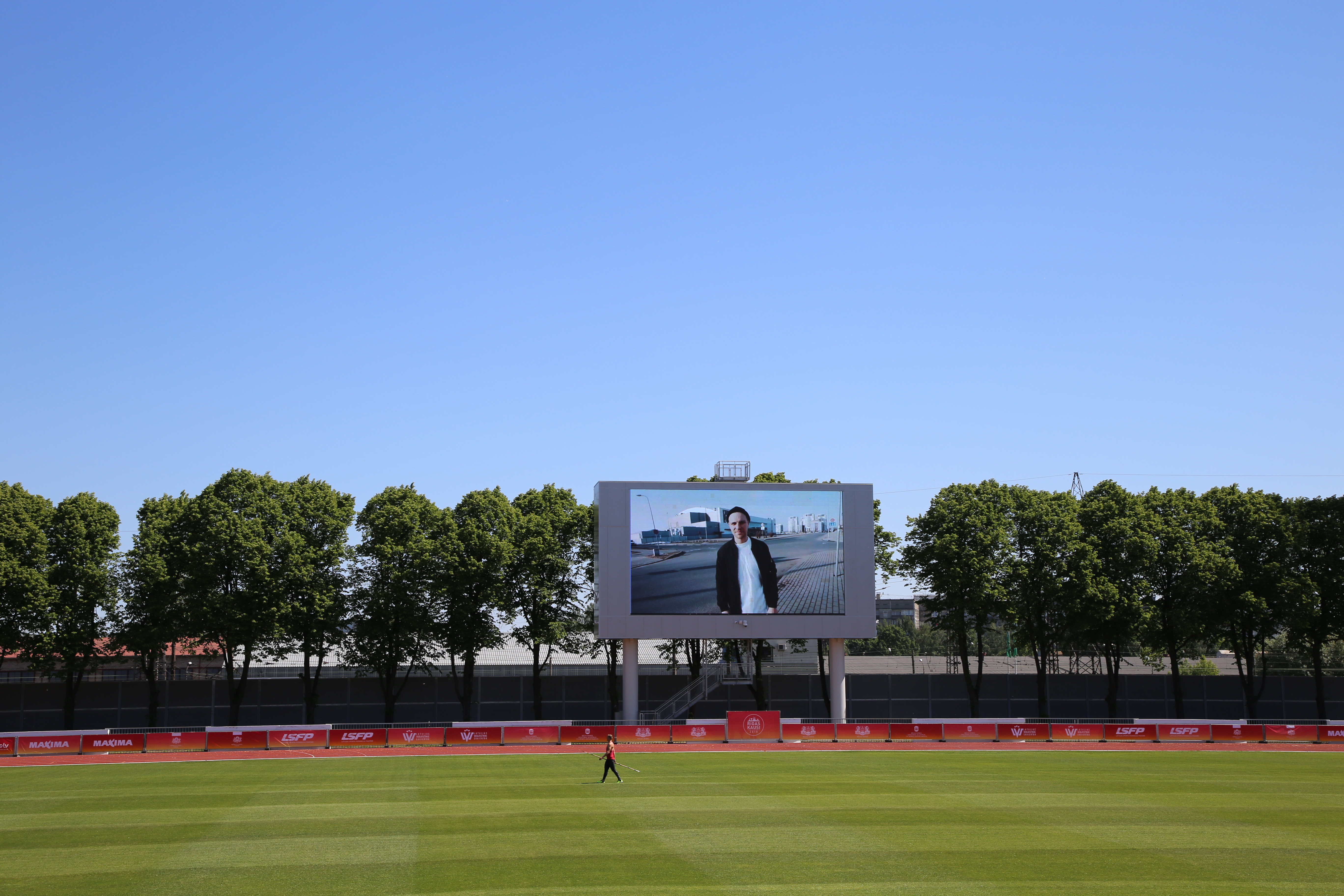 Foto:  Daugavas stadiona tablo, uz kura redzams reperis Ozols, cilvēks futbola laukuma zaļajā zonā nūjo