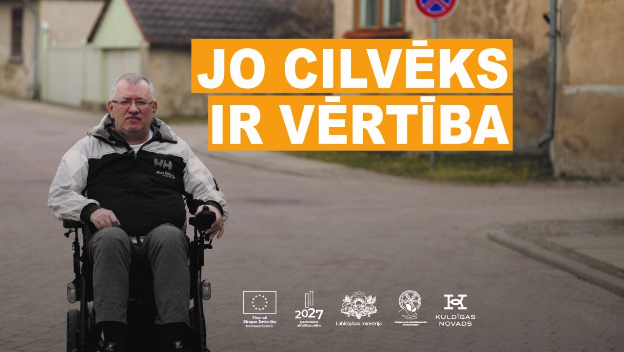 Bruģēta vecpilsētas iela. Vīrietis ap 50 gadiem, īsiem, sirmiem matiem brillītēs un pelēkā rudens vējjakā sēž elektriskajā ratiņkrēslā. Uzraksts 