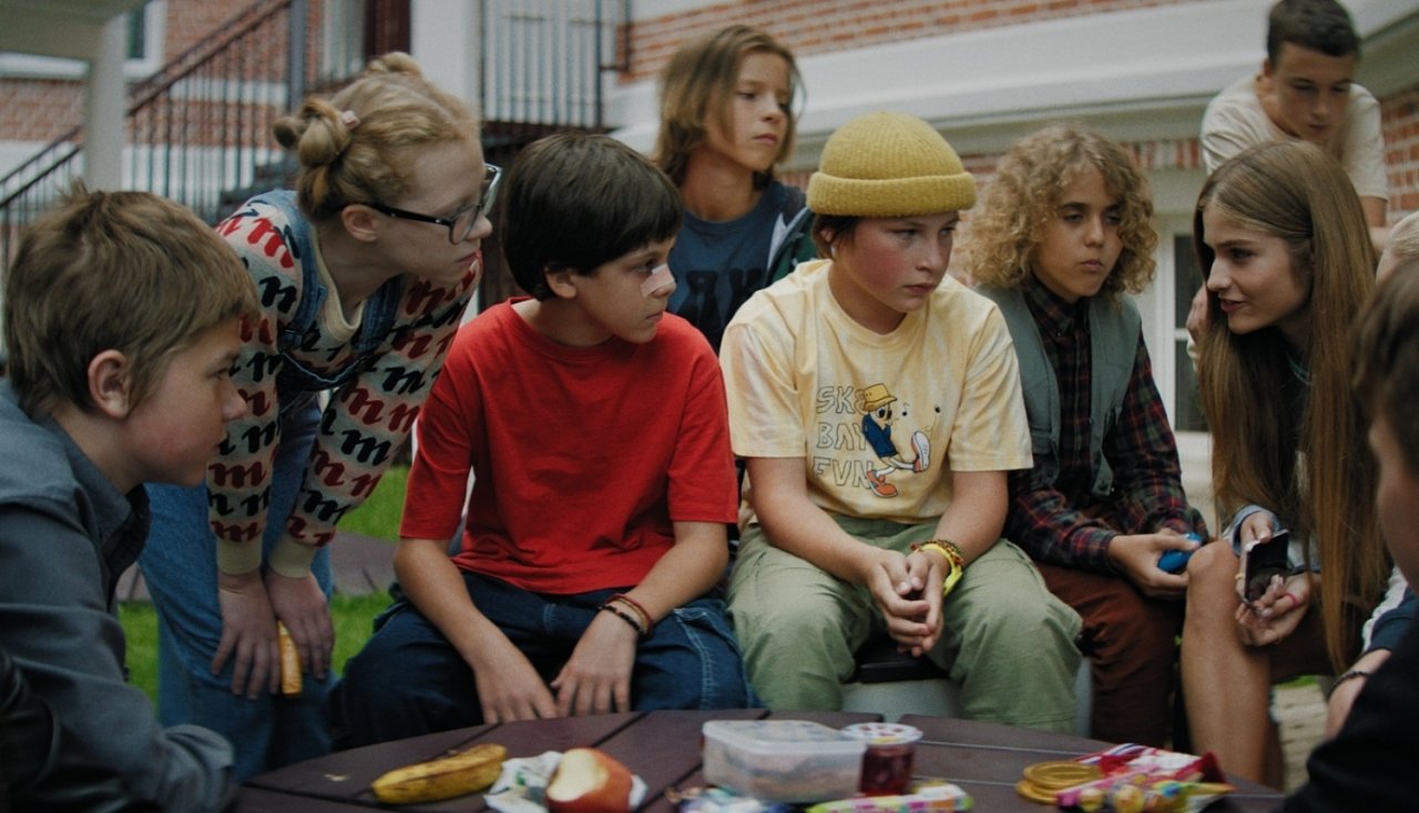 A shot from the film shows children sitting in a group and talking