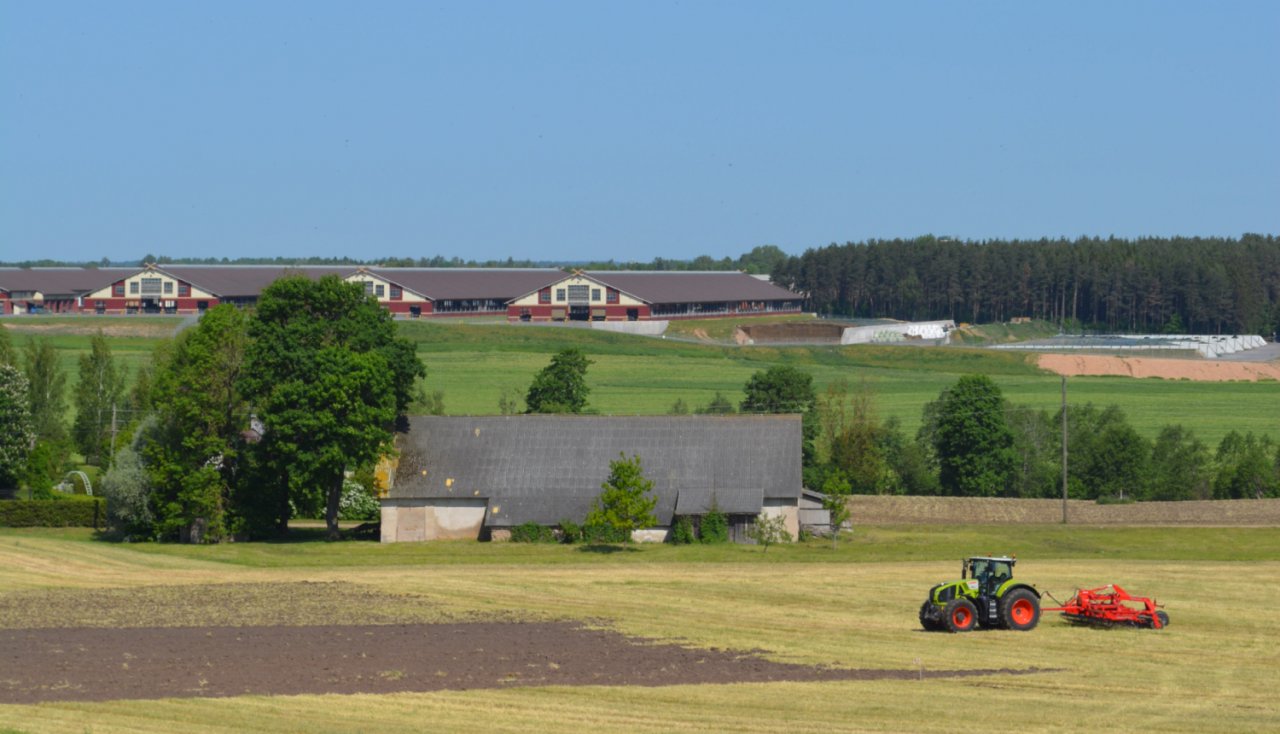 Skats uz lauku aimniecību - priekšpānā traktos apstrādā zemi, aiz tā veca saimniuecības ēka, pakalnā augšā jaunas fermas ēkas