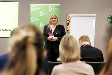 Sieviete stāv auditorijas priekšā un kaut ko stāsta