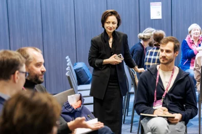 Olga Kazāka, pasākuma vadītāja, stāv un vēro kā cilvēki darba grupās strādā