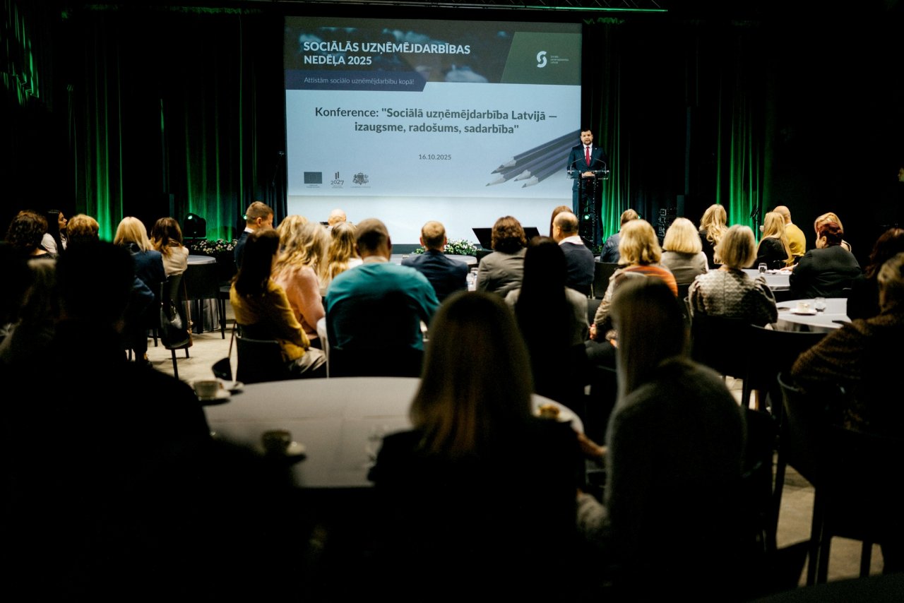 Konferences telpa. Auditorija (cilvēki virs 30) sēž pie galdiņiem. Uz skatuves liels ekrāns un labklājības ministrs sniedz runu