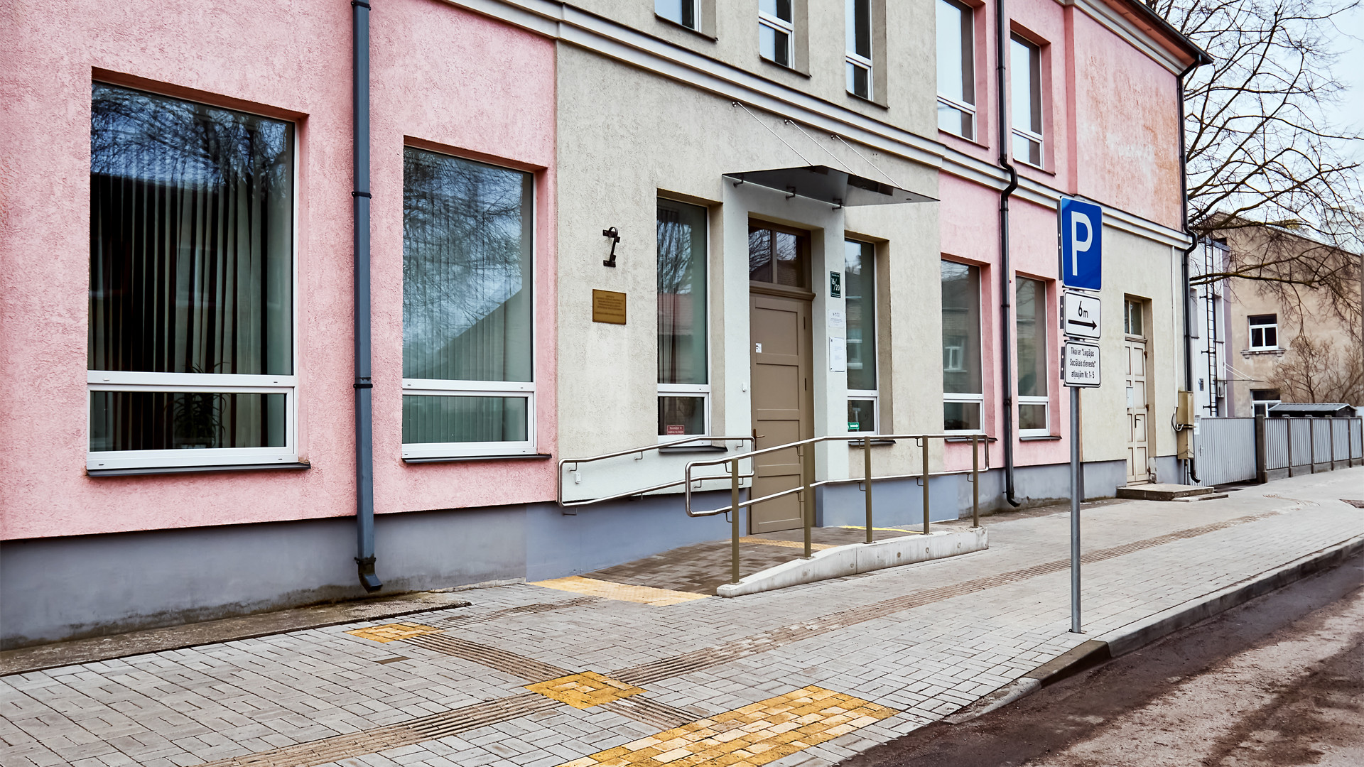 Divstāvīgas mājas fasāde. Priekšā neliels bruģēts panduss ar fasādes durvīm. Bruģēta ietve ar takstilajām līnijām. Asfaltēts ceļš. Stāvlaukuma zīme troturāra malā.