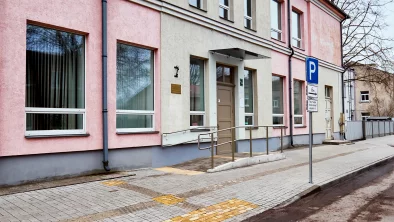 Divstāvīgas mājas fasāde. Priekšā neliels bruģēts panduss ar fasādes durvīm. Bruģēta ietve ar takstilajām līnijām. Asfaltēts ceļš. Stāvlaukuma zīme troturāra malā.