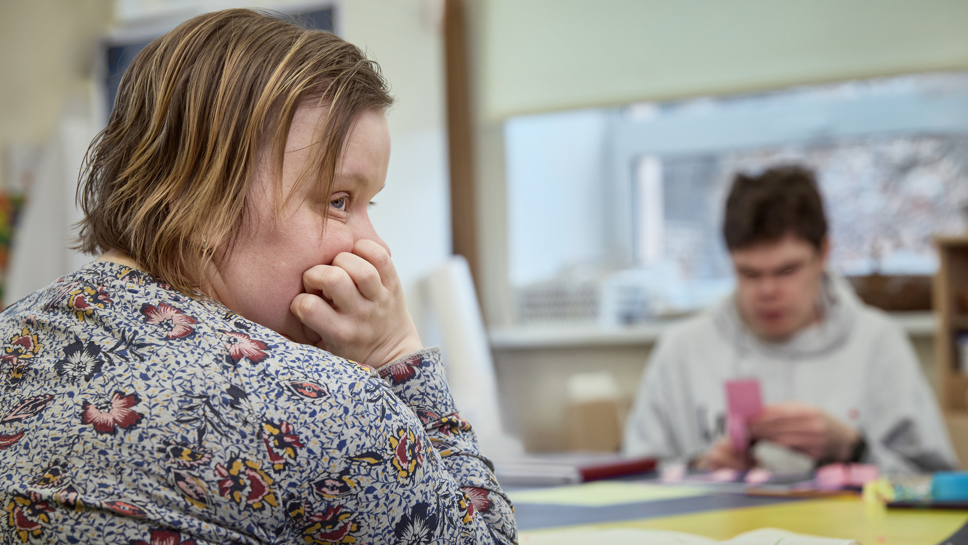 Domīga jauna sieviete atspiedusi galvu uz rokām un rokas atbalstijusi uz galda kaut ko vēro. Fonā jauns vīrietis nodarbojas papīra griešanu