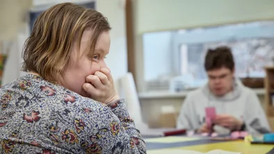 Domīga jauna sieviete atspiedusi galvu uz rokām un rokas atbalstijusi uz galda kaut ko vēro. Fonā jauns vīrietis nodarbojas papīra griešanu