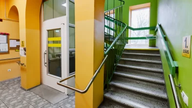 Kāpņu telpa. Sienas krāsotas zaļā un dzeltenā krāsā, tumši pelēka flīzēta grīda, tumši pelēkas kāpnes. Margas apbās trepju pusēs. Lifta durvis kreisajā pusē.