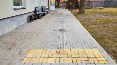 Iekšpagalms. Kreisajā pusē mājas siena ar diviem krēsliem, labajā zāliens ar diviem lieliem kokiem, šūpolēm un brūnu koka žogu. Pa vidu plata bruģēta ietve ar taktilo joslu.