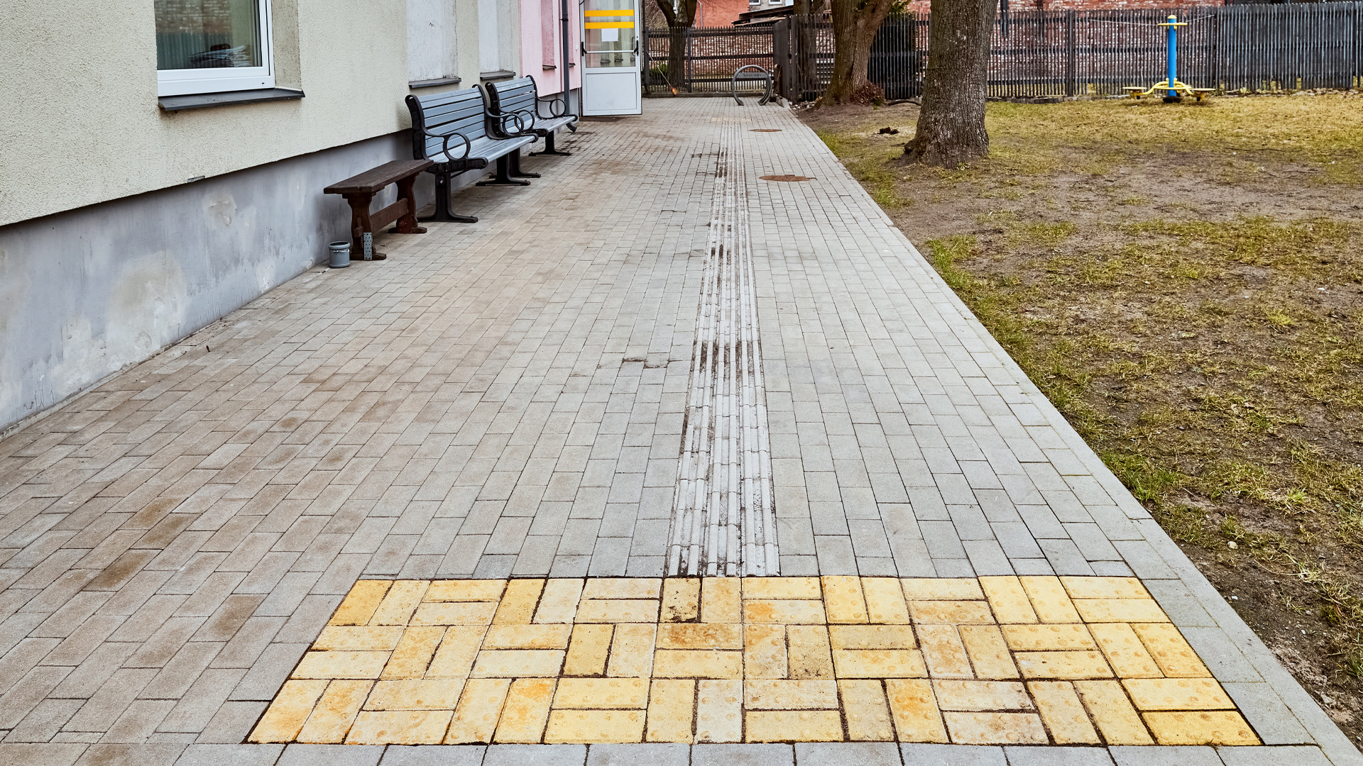 Iekšpagalms. Kreisajā pusē mājas siena ar diviem krēsliem, labajā zāliens ar diviem lieliem kokiem, šūpolēm un brūnu koka žogu. Pa vidu plata bruģēta ietve ar taktilo joslu.