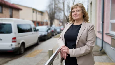 Sieviete ap 50 gadiem ar bondiem matiem, melnā blūzē un bēšīgu žaketi stāv pie mājas ietves pusē