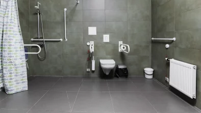Cilvēkiem ar invaliditāti pilnībā pielāgota tualete ar dušu. Tumši pelēka flīzēta grīda. Tumši zaļas flīzētas sienas
