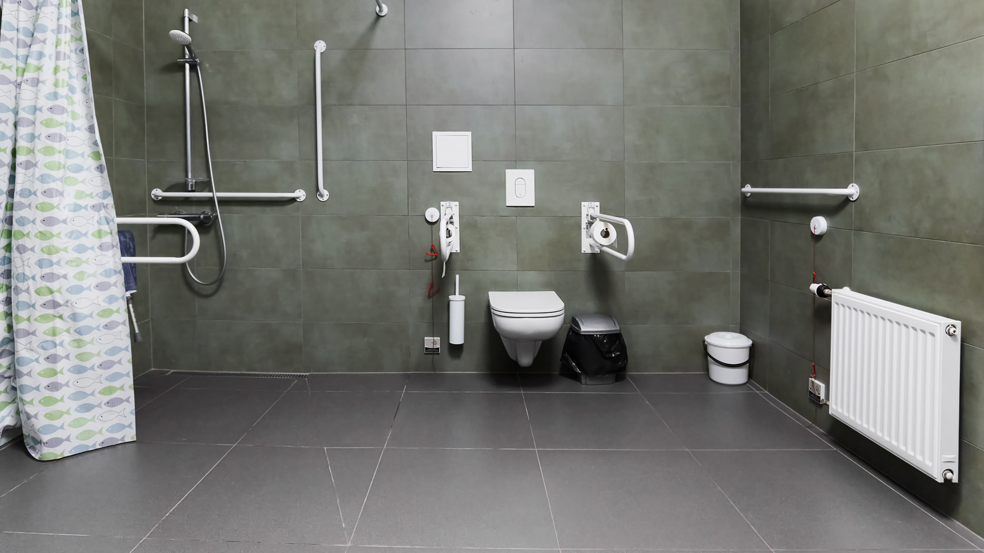 Cilvēkiem ar invaliditāti pilnībā pielāgota tualete ar dušu. Tumši pelēka flīzēta grīda. Tumši zaļas flīzētas sienas