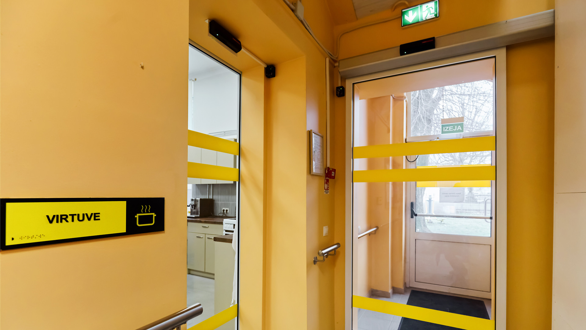 Neliels koridoris ar vairākām stiklotām un dzeltenām sienām. Pa perimetru roku balsti un kreisajā pusē durvju norāde ar uzrakstu 