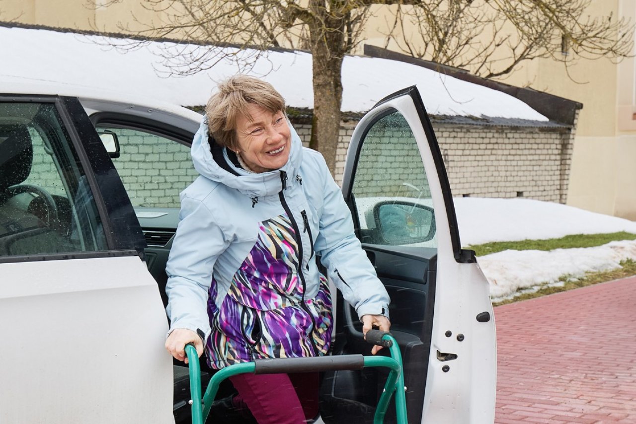 Sievieta ap 50 gadu vecumam smaidot kāpj ārā no balta auto.