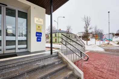 Ēkas pilsētā ārpuse ziemā. Redzama iela, sarkans gājēju bruģis, kāpnes ar margām un ieejas durvis.