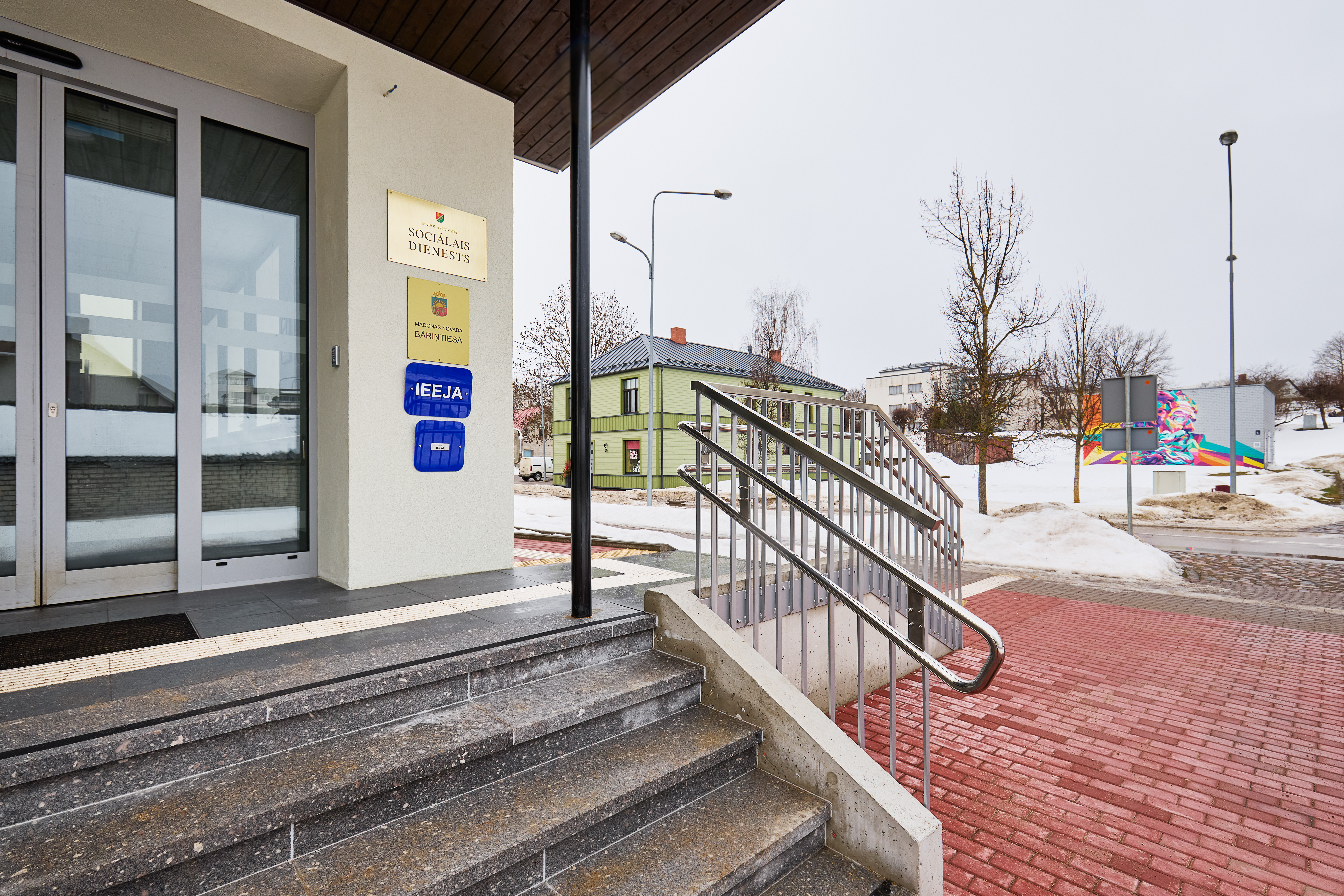 Ēkas pilsētā ārpuse ziemā. Redzama iela, sarkans gājēju bruģis, kāpnes ar margām un ieejas durvis.