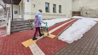 Ēkas ārpuse, fonā kāpnes un alternatīvs bruģēts celiņš, kas pilda pandusa funkciju. Sieviete ar skaigulīti iet pa burģēto celiņu