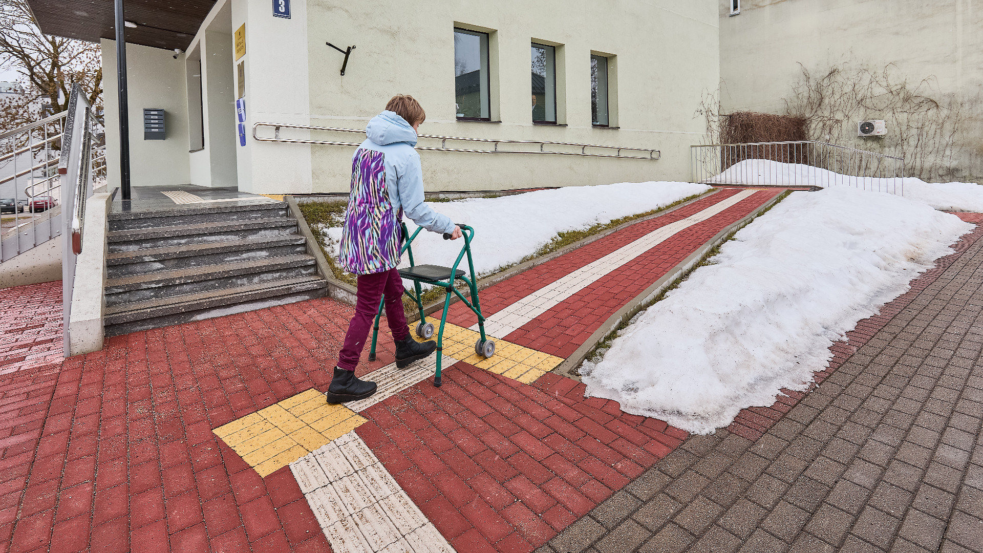 Ēkas ārpuse, fonā kāpnes un alternatīvs bruģēts celiņš, kas pilda pandusa funkciju. Sieviete ar skaigulīti iet pa burģēto celiņu