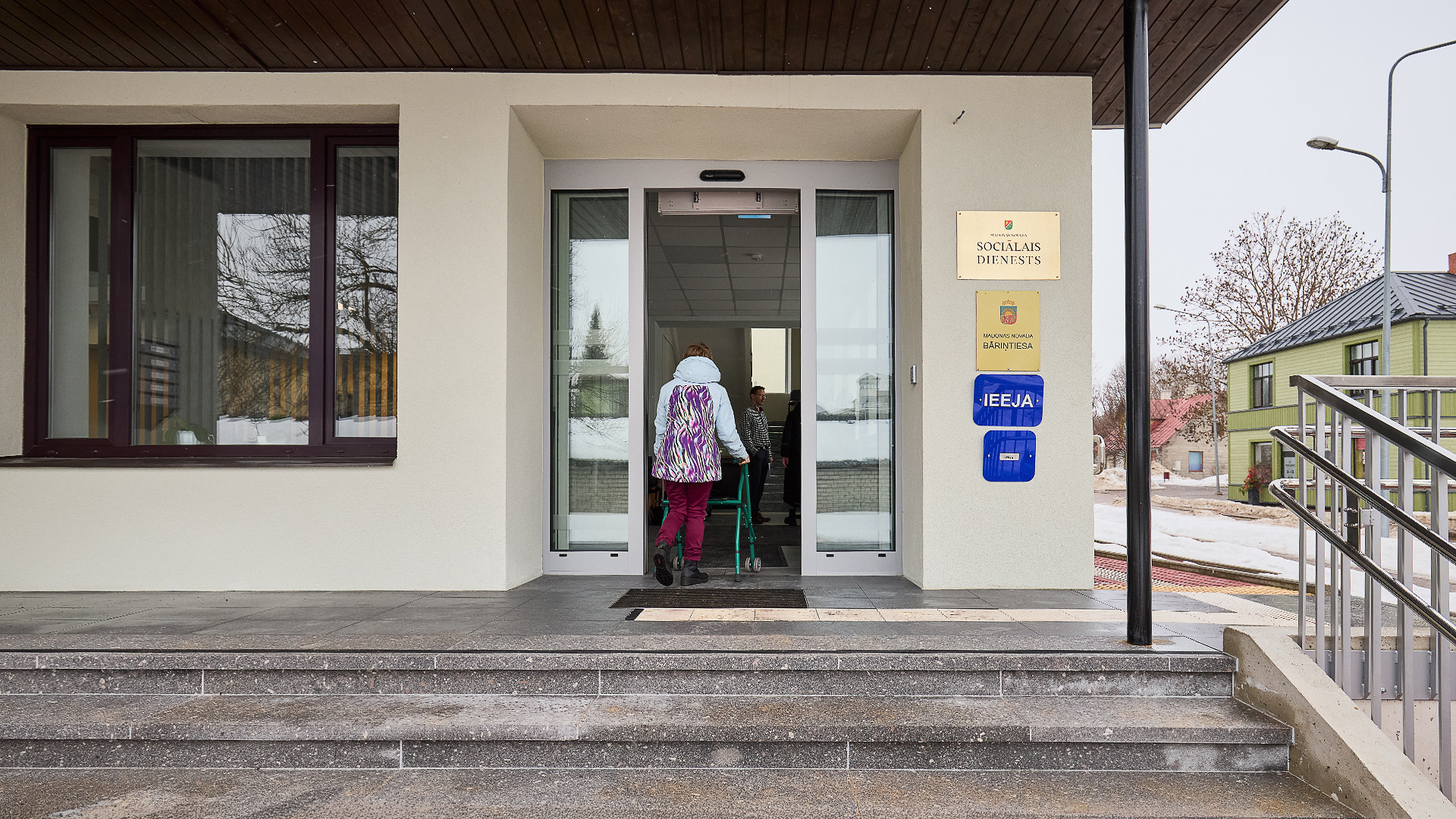 Sieviete ar staigulīti iet iekšā pa ēkas ieeju ar automātiski veramajām durvīm.