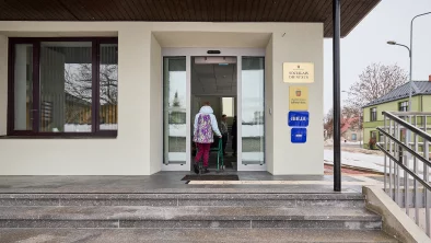 Sieviete ar staigulīti iet iekšā pa ēkas ieeju ar automātiski veramajām durvīm.