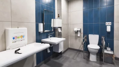 Tualetes telpa. Divu veida sienu flīzējums: balts un tumši zils. Pelēka grīda. Redzams zīdaņu pārsienamais galdiņš, izlietene ar spoguli un dvieļu turētāju, tualetes pods ar balstiem cilvēkiem ar invaliditāti.