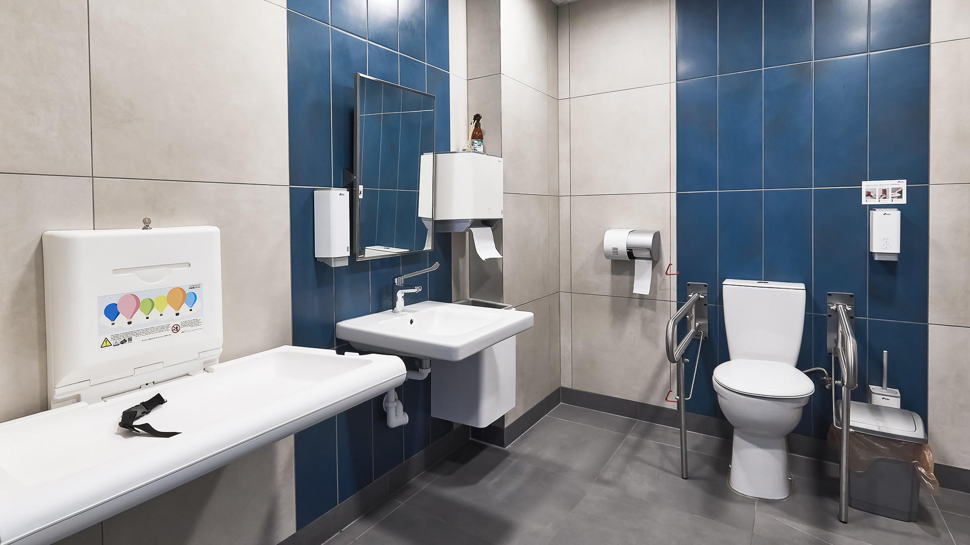 Tualetes telpa. Divu veida sienu flīzējums: balts un tumši zils. Pelēka grīda. Redzams zīdaņu pārsienamais galdiņš, izlietene ar spoguli un dvieļu turētāju, tualetes pods ar balstiem cilvēkiem ar invaliditāti.