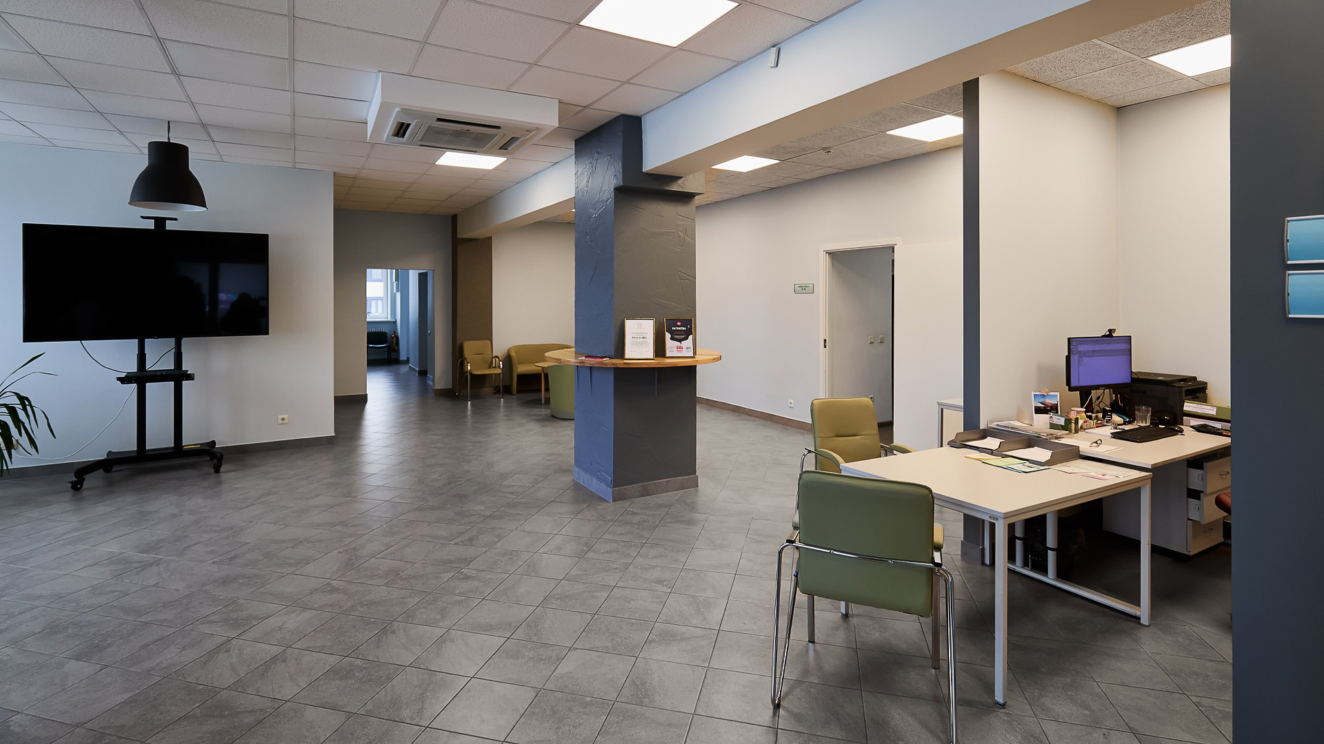 Plaša, gaiši pelēka iekštelpa. Uz grīdas tumši pelēkas flīzes.  Fonā biroja galds ar datoru un diviem krēsliem.