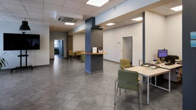 Plaša, gaiši pelēka iekštelpa. Uz grīdas tumši pelēkas flīzes.  Fonā biroja galds ar datoru un diviem krēsliem.