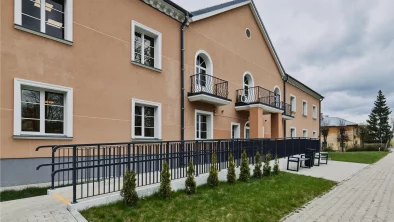 Divstāvīgas oranžas ēkas ar baltiem logu rāmjiem un melnām margām fasāde. Pie fasādes jauns bruģis, zāliens un divi soliņi