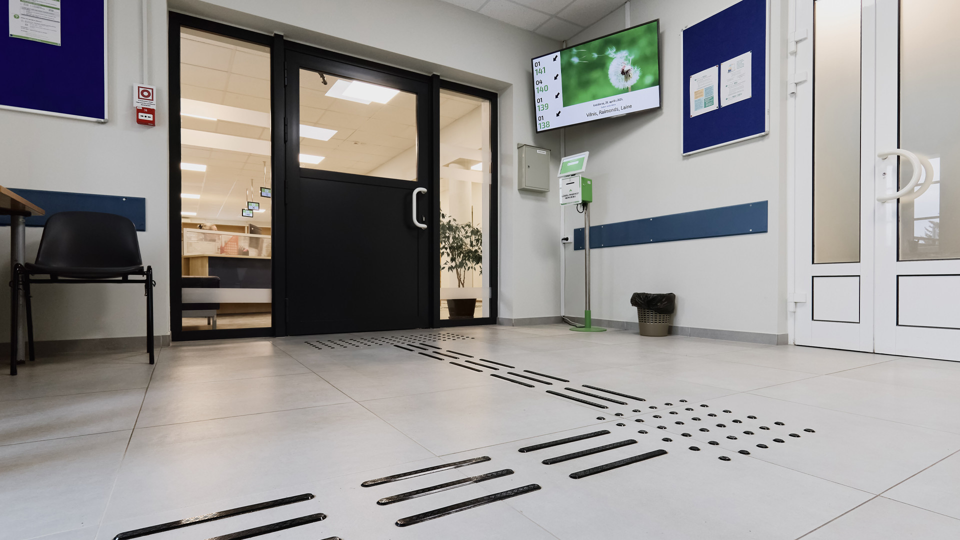 Uzgaidāmā telpa ar platām durvīm uz nākamo telpu, lielu informatīvo TV un aparātu klienta kārtas numuriņa izņemšanai