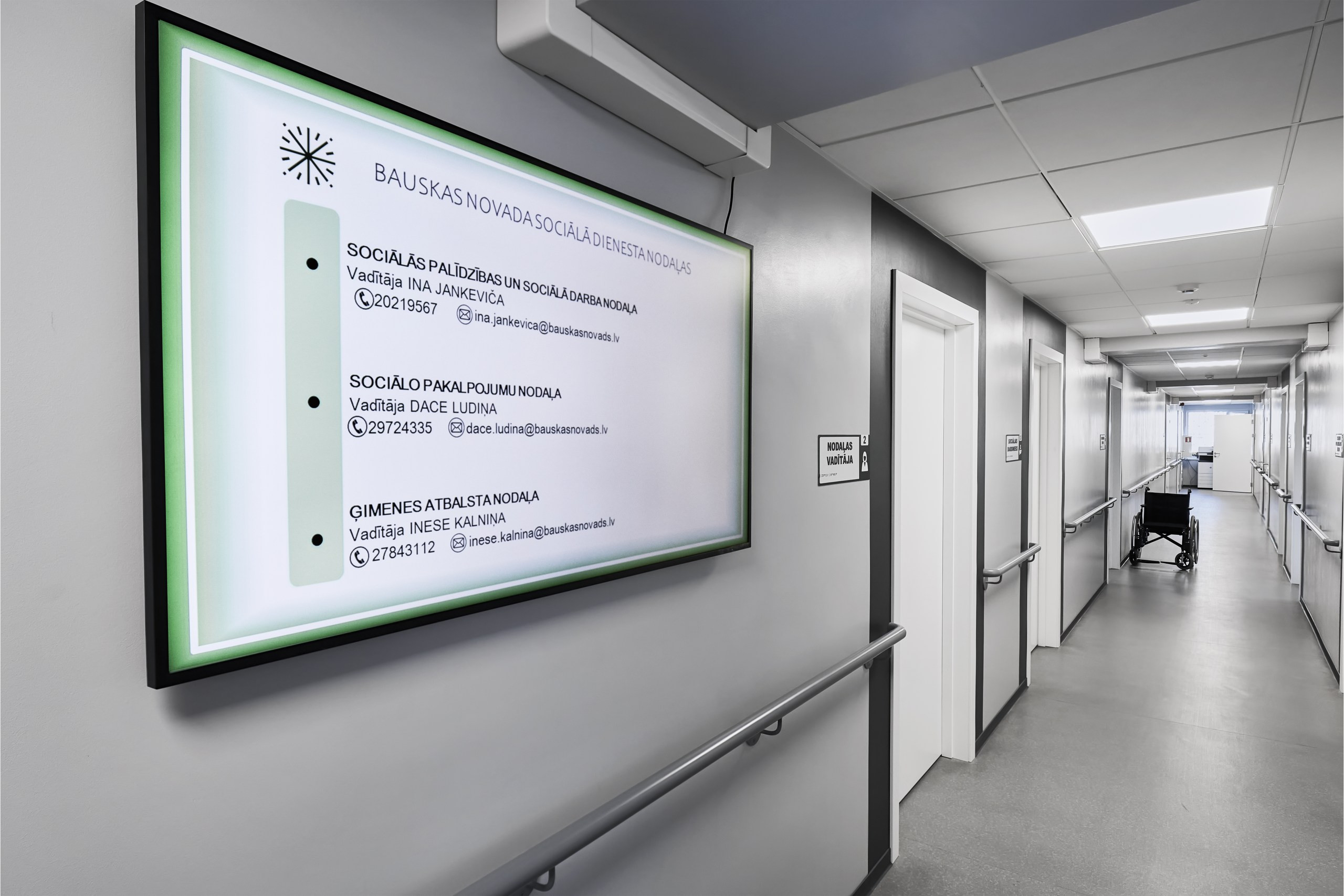 Pelēks koridoris ar daudz durvīm. Pie sienām margas. Uz sienas liels monitors ar informāciju par sociālo dienestu. Koridora vidū ratiņkrēsls.