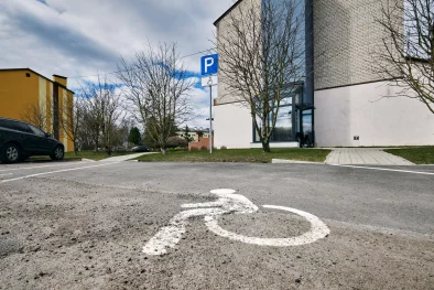 Ēkas ārpuse. Skats no ārpagalma cilvēku ar invaliditati stāvlaukuma uz stiklotu ieeju ēkā.
